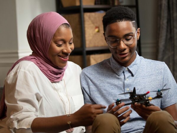 A Black woman in a hijab looks at a small drone with her teenage son