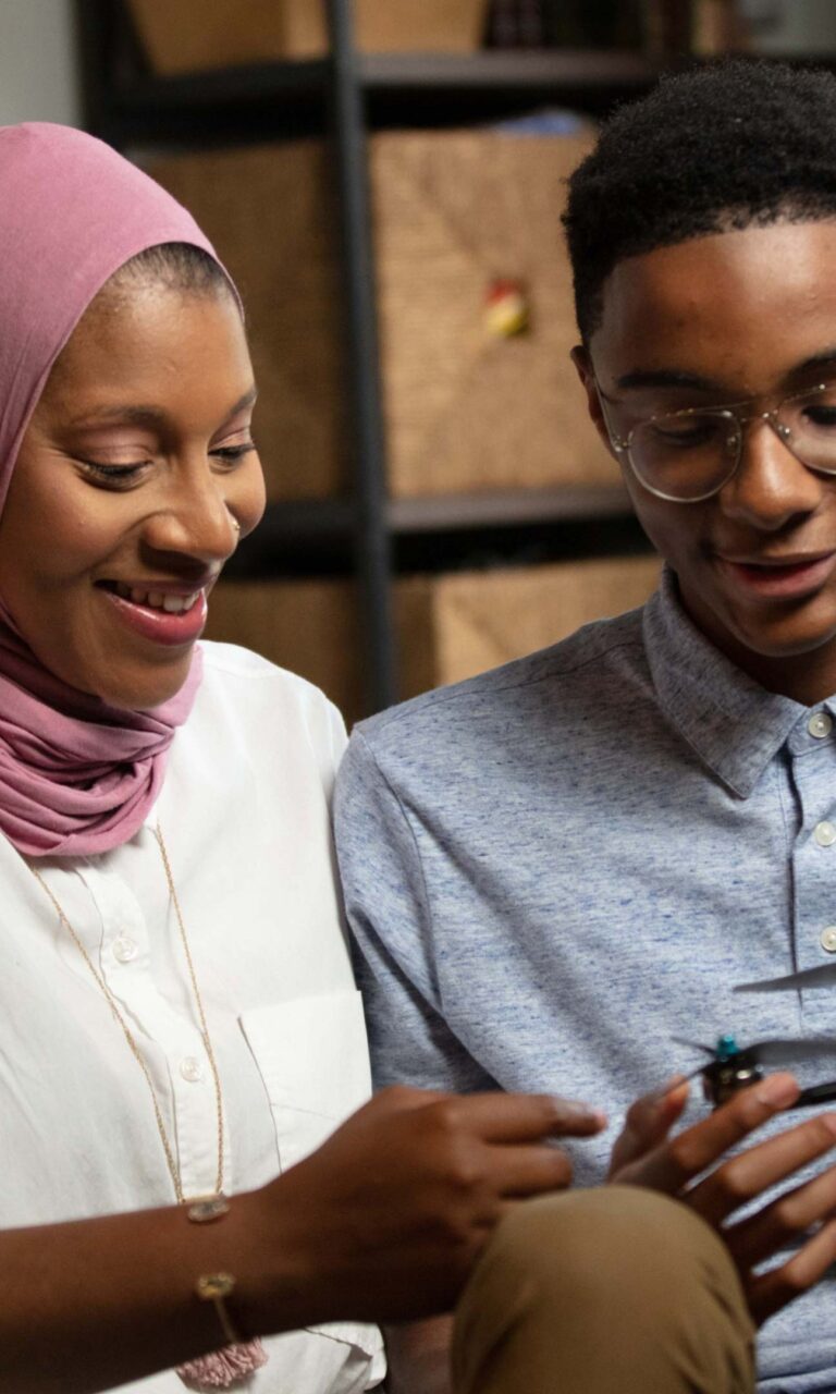A Black woman in a hijab looks at a small drone with her teenage son