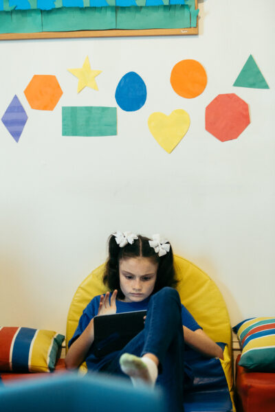 young caucasian girl with brown hair in pigtails with white bows sitting in a yellow chair playing on ipad tablet
