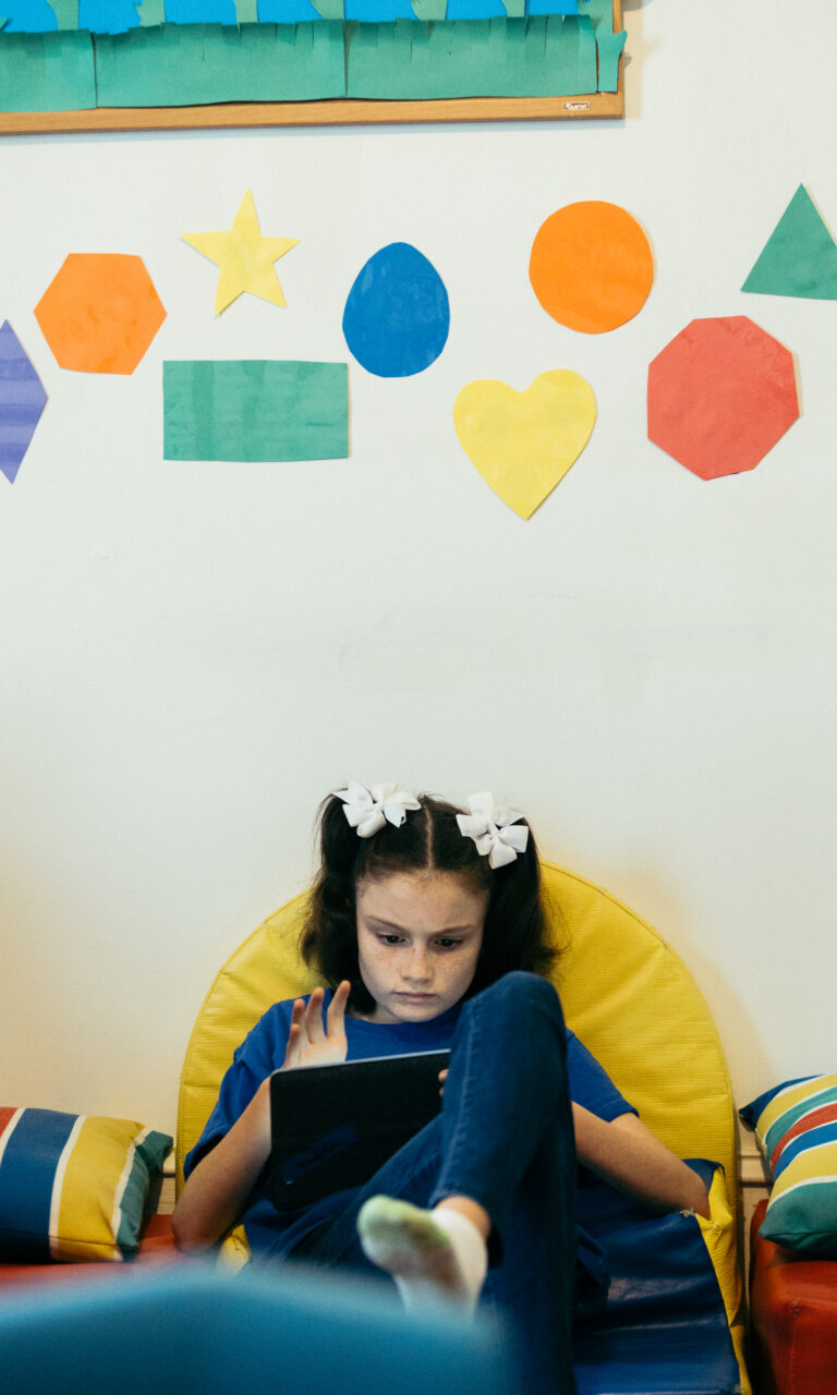 young caucasian girl with brown hair in pigtails with white bows sitting in a yellow chair playing on ipad tablet