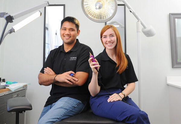 Ryan and Hannah Maruyama of The Degree Free Podcast sitting in their tattoo studio in matching black t-shirts.