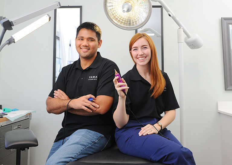 Ryan and Hannah Maruyama of The Degree Free Podcast sitting in their tattoo studio in matching black t-shirts.