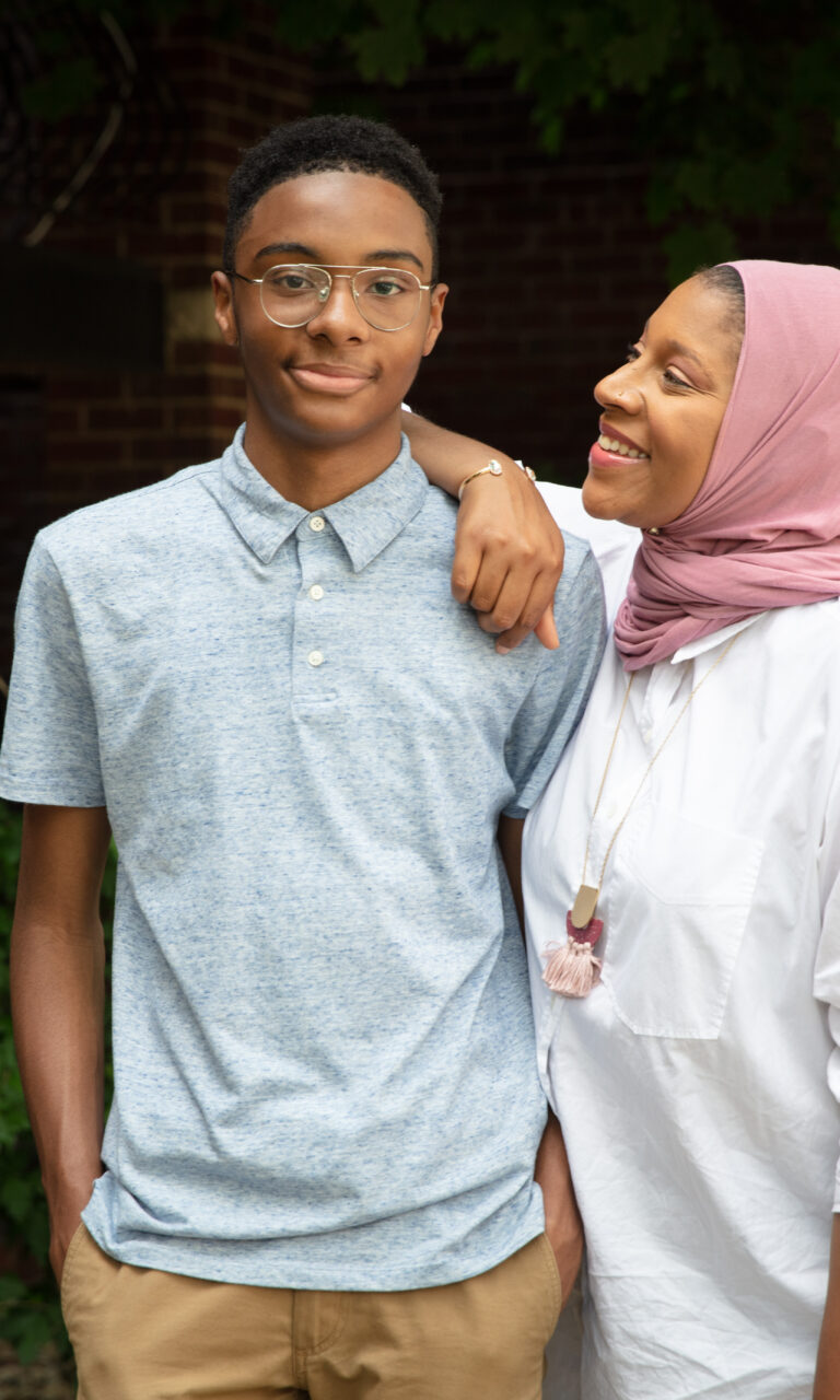 A mother in a hijab with her arm on her teenage son's shoulder.