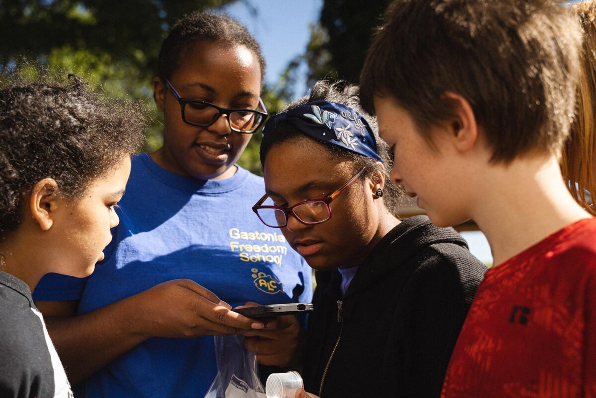 A Black woman takes a picture of something with her cellphone. Three young children are gathered watching