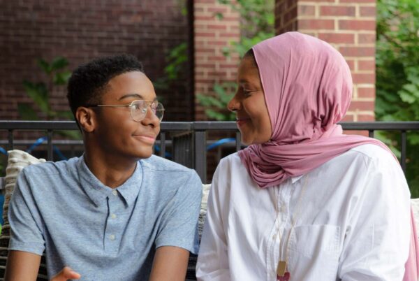 A mother in a hijab looks at her teenage son in glasses, and they're both smiling.