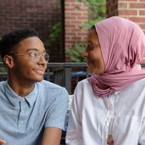A mother in a hijab looks at her teenage son in glasses, and they're both smiling.