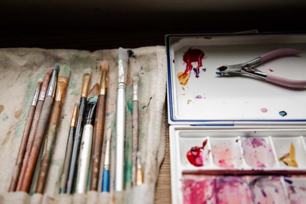 A bunch of paint brushes on lying out next to a tray filled with variously hued pink paints.