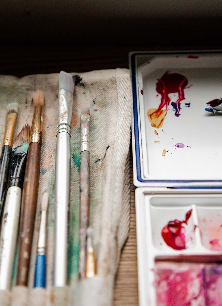A bunch of paint brushes on lying out next to a tray filled with variously hued pink paints.
