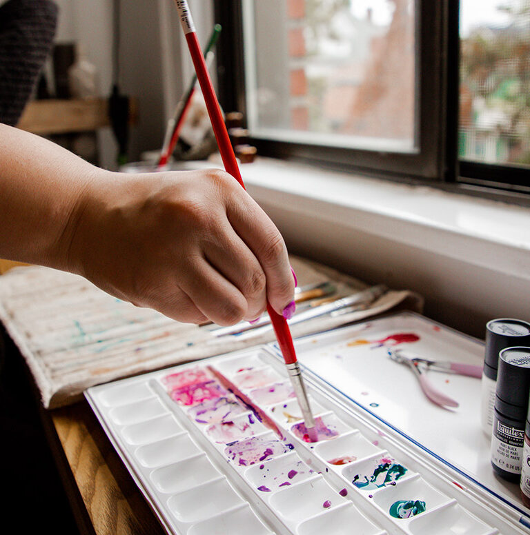 Sarah holds a paintbrush to a tray, getting a pinkish red color on the brush.