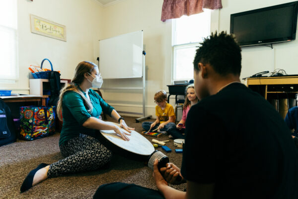 A teacher in a classroom banging on a drum while the kids gather around her in a semi-circle playing their own percussion.