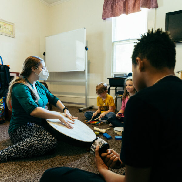 A teacher in a classroom banging on a drum while the kids gather around her in a semi-circle playing their own percussion.