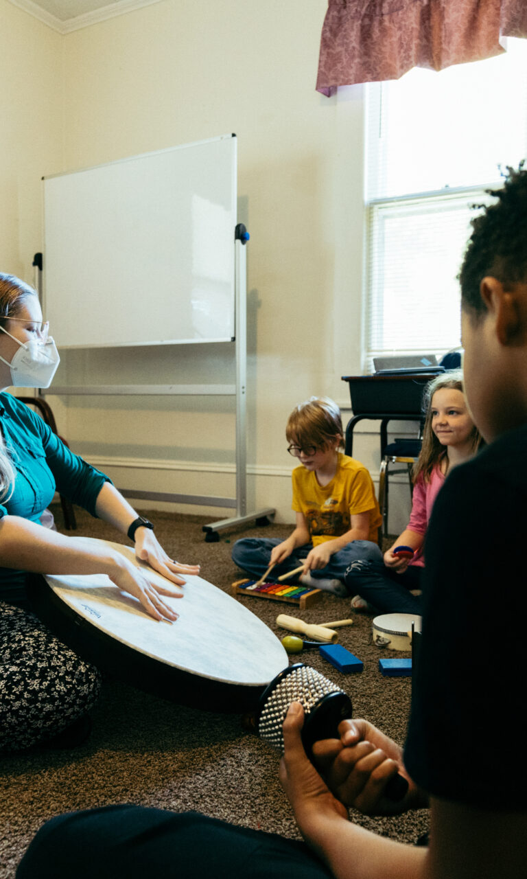 A teacher in a classroom banging on a drum while the kids gather around her in a semi-circle playing their own percussion.