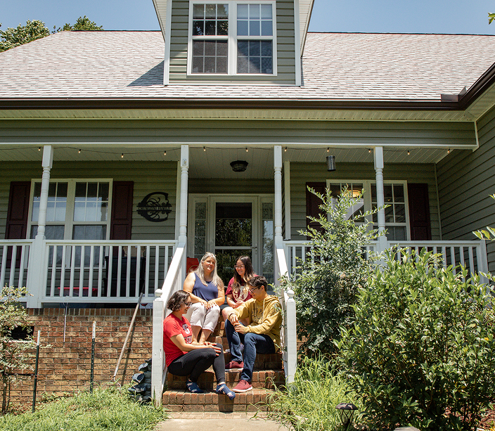 A family of 4 sitting on the front steps of their porch in sunny weather.