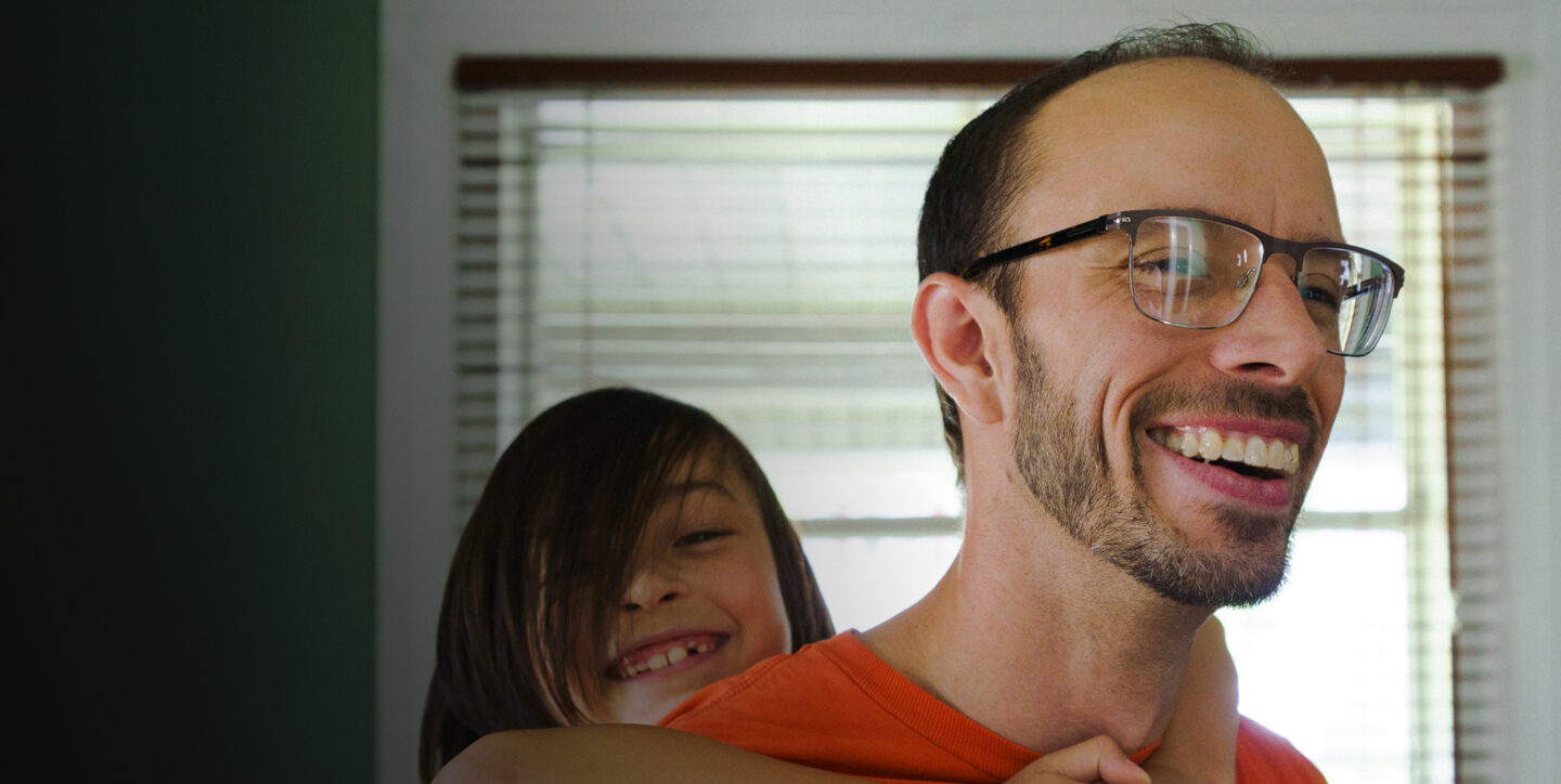 A son, Kai, rides piggy back on his Dad's back. Both have wide smiles.