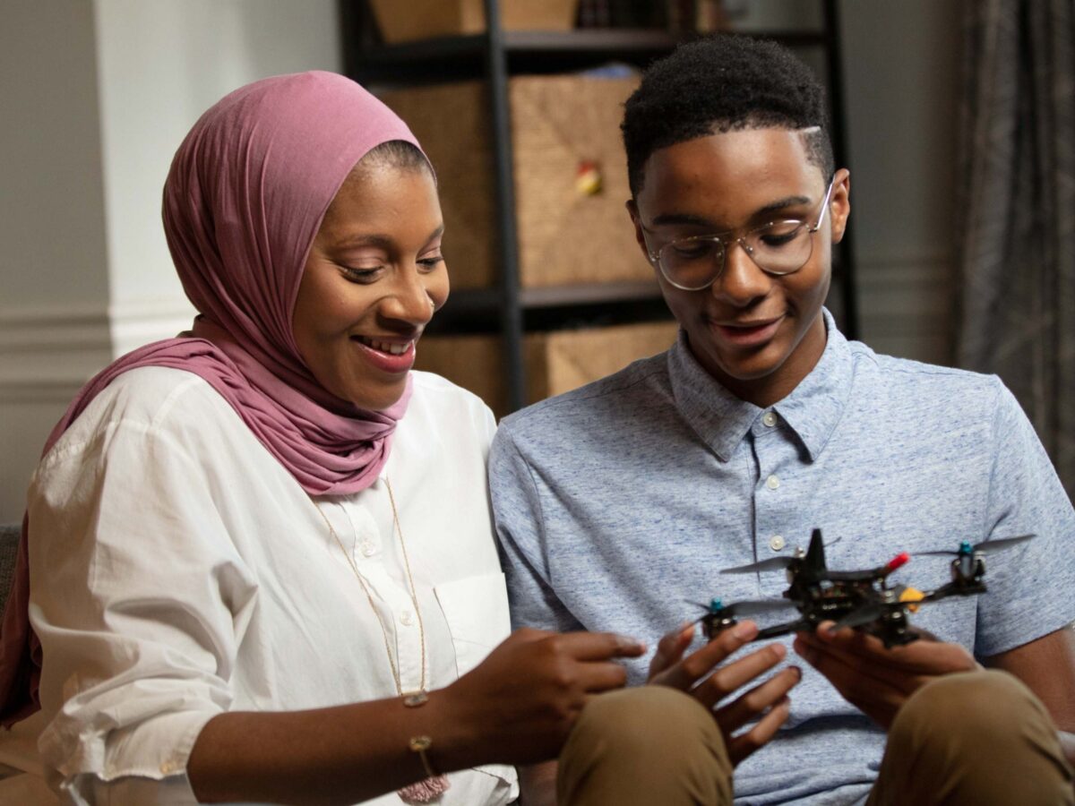 A Black woman in a hijab looks at a small drone with her teenage son