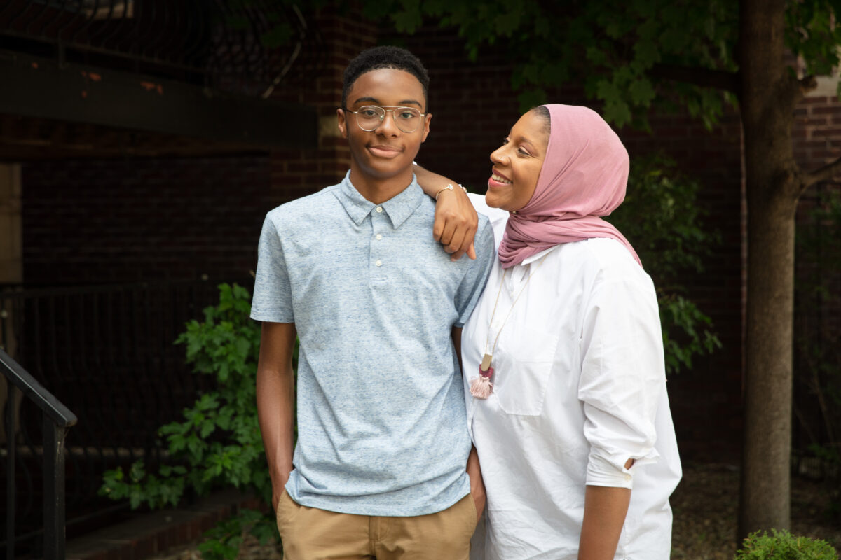 A mother in a hijab with her arm on her teenage son's shoulder.