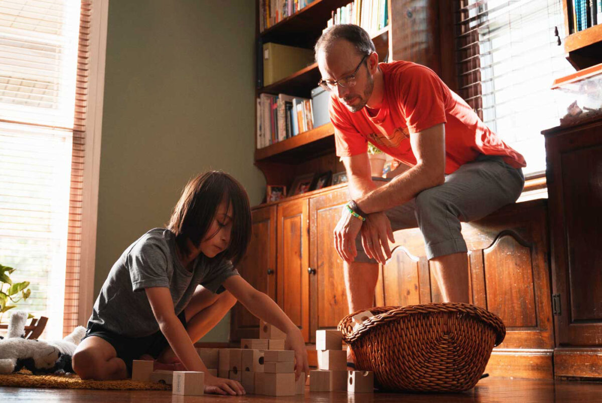 A kid playing with big wooden blocks while his father looks on.